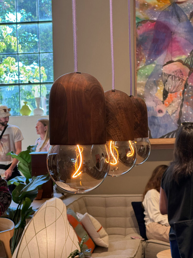 Three Light Bean pendant lights by Katerina Kopytina, with wooden tops and exposed filaments, hang above a seating area in a room with several people and a colorful wall painting.
