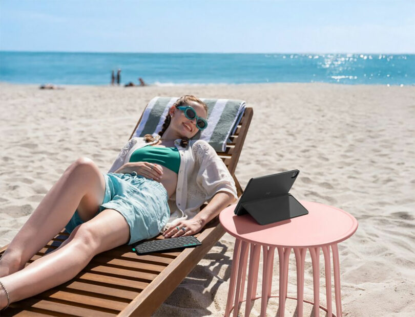 A woman relaxes on a lounge chair at the beach, her tablet in a Flip Folio case and keyboard set on a small pink table beside her. The ocean and blue sky create a serene backdrop.