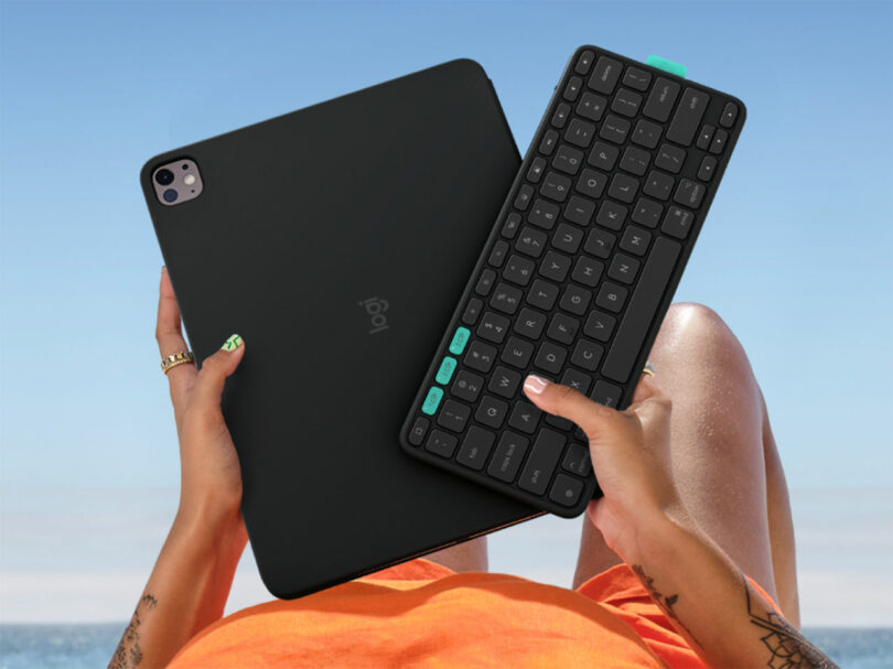 A person holds a tablet with a Flip Folio black case in one hand and a wireless keyboard with teal keys in the other, while sitting outdoors with the sky and sea in the background.