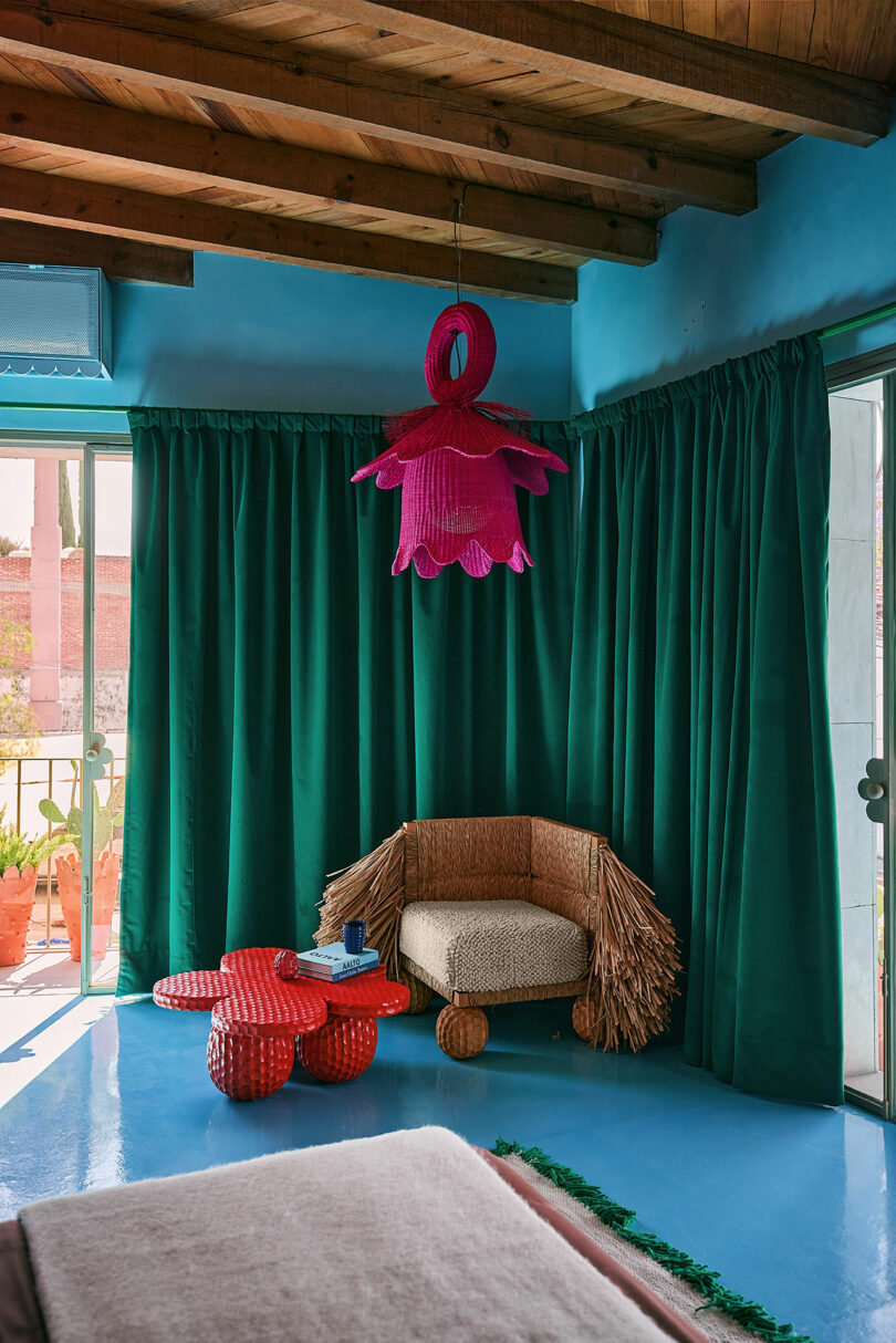 Room with blue walls and floor, green curtains, woven chairs with frigged hands, red sculptural coffee table and large pink ceiling shaped in the shape of flowers.