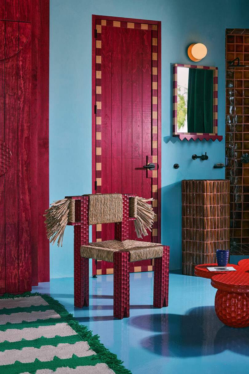 Room with blue walls, red and tan doors, woven chair with ropes details, round red table, wall mirror and green.