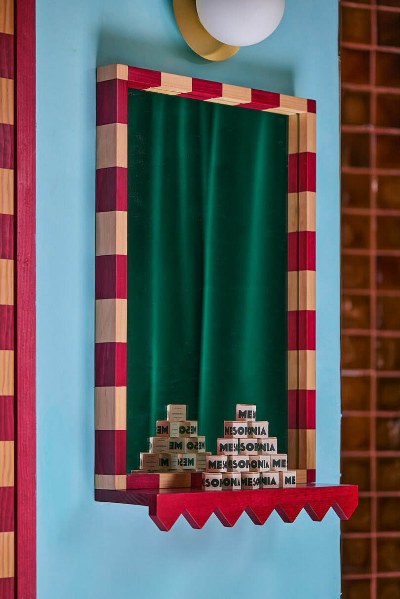 Rectangular mirror with red and beige Carized frame that reflects wooden blocks on the spelling of the shelf "Flood" and "Me" in different arrangements.