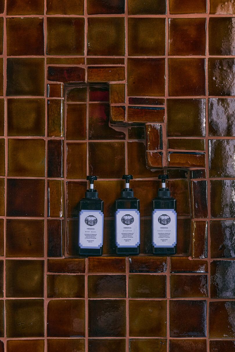 Three bottles with a dark pump with white labels mounted on a brown paved wall, arranged vertically in the recessed tiled section.