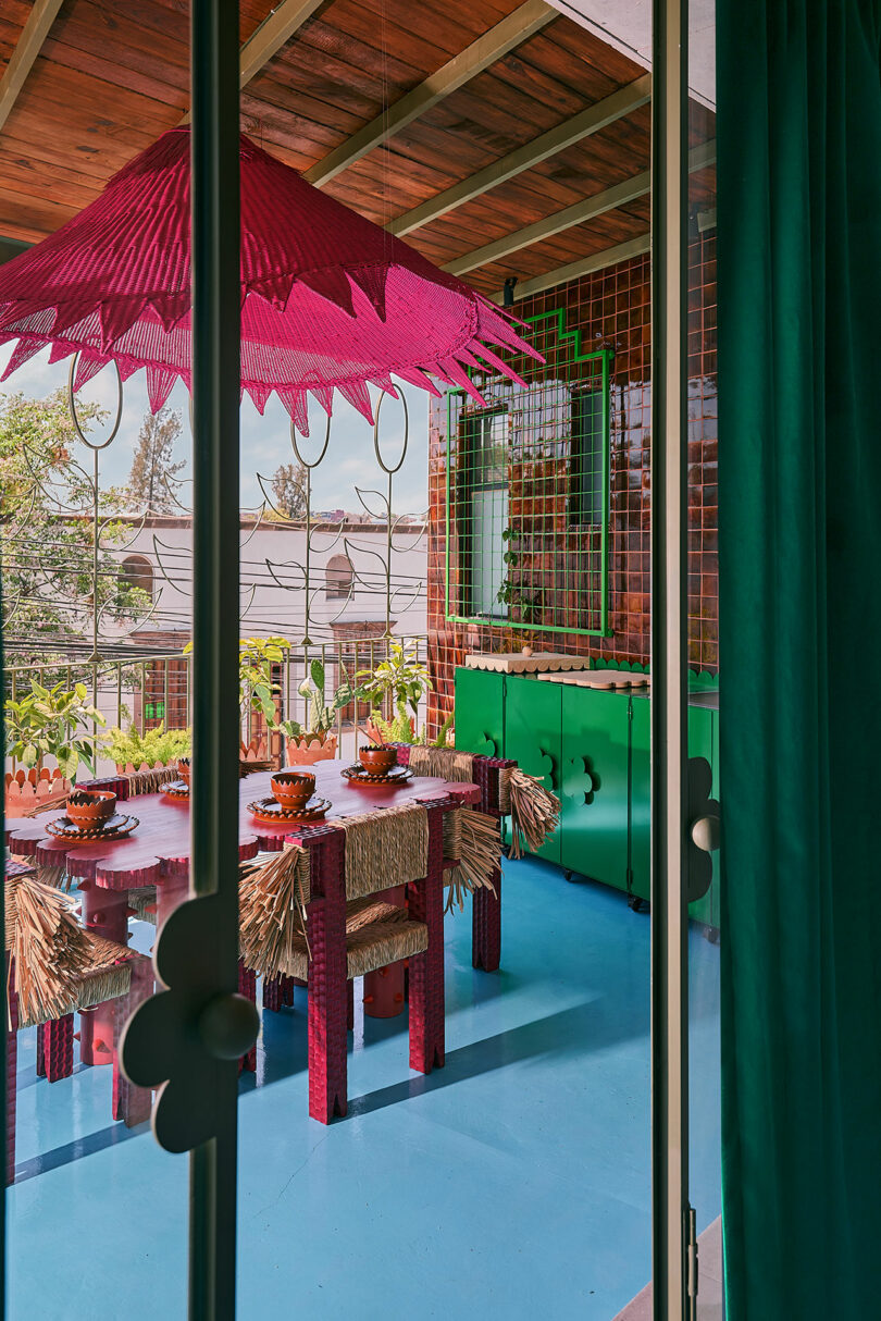 A colorful dining area with a pink umbrella, straw chairs, green cabinets and blue floors, viewed through the glass door.