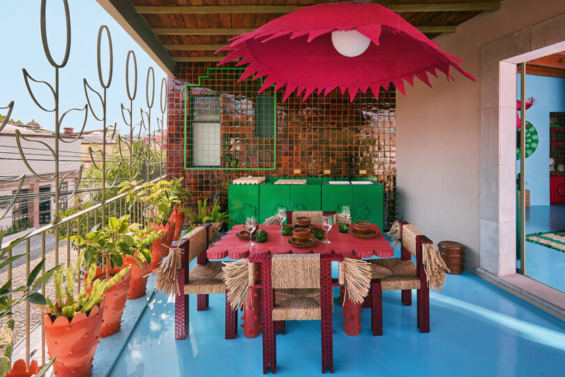 Colorful dining area outdoors with pink umbrella, straw chairs, terracotta codes, mosaic tiles and green cabinets on the blue floor.