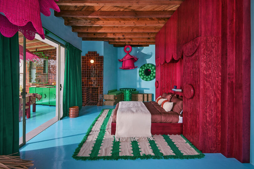 Bedroom with blue floor, red wood wall, green and white mat, brown bed and open glass door leading to a neighboring room with green decor and wooden ceilings.