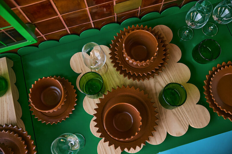 Table Set with brown ceramic vessels, green glass cups, wine glasses and wooden shrimps on a green surface, with a brown tiled background in the background.