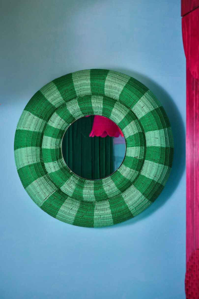 The round mirror with a green-checked woven frame hangs on the light blue wall with pink and green curtains partially visible in reflection.