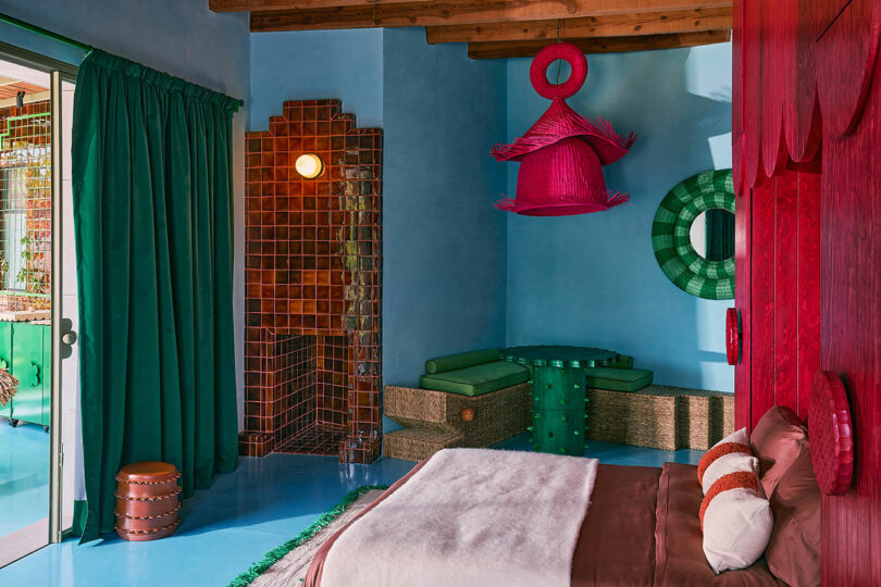 Colorful bedroom with blue walls, red accent, bed with brown and white bedding, green angled sedentic space and sliding glass door with green curtains.