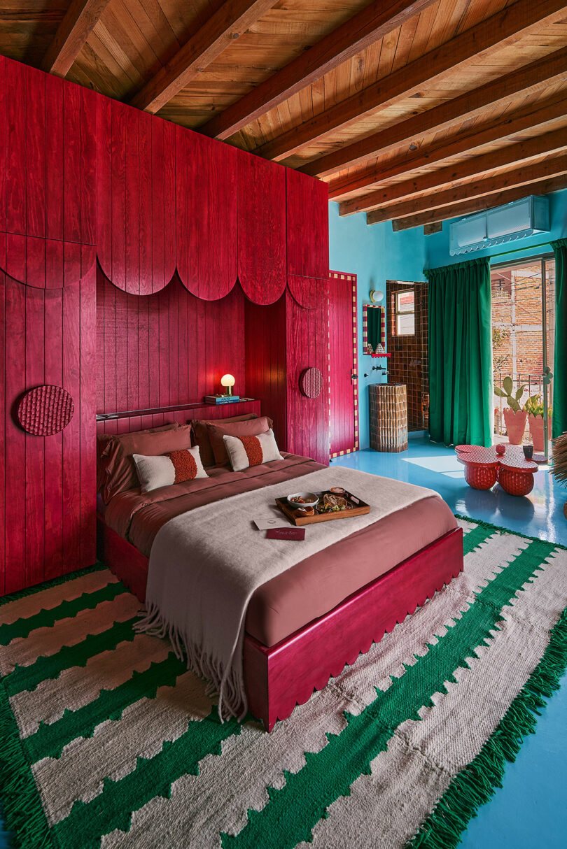 Bedroom with brave red emphasis and bed, green curtains, green and white covers, blue colored floor and wooden ceiling beams. The natural light enters the window.