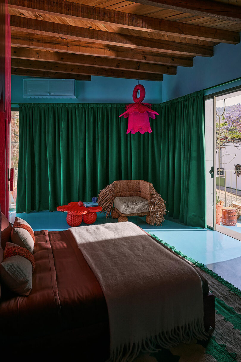 Bedroom with wooden ceiling, green curtains, brown bed, unique pink ceiling lamp, knitted table and red tables, with sunlight that passed through the sliding glass door.
