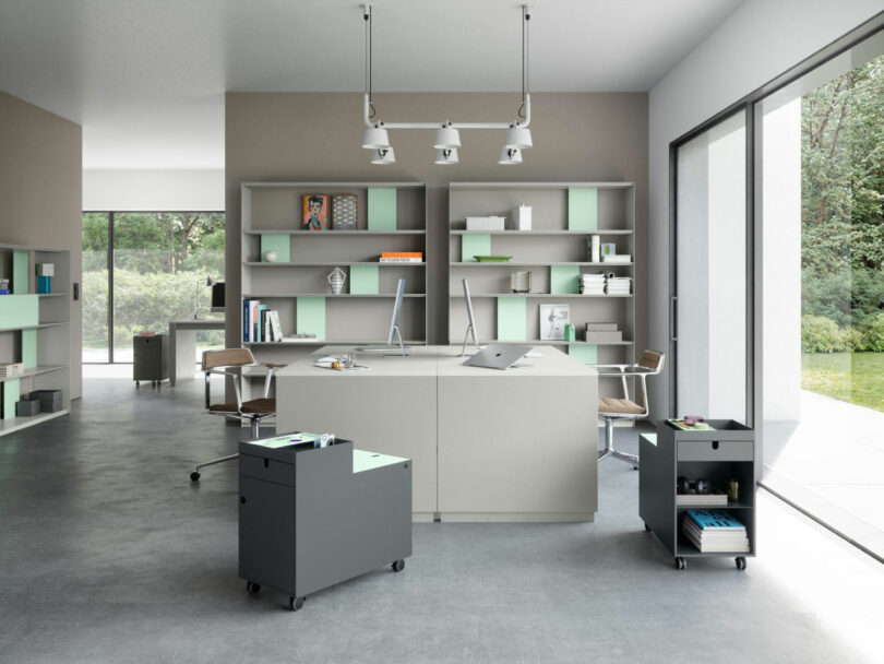 Modern office space with large windows, minimalist desks, open shelving, and rolling storage units on a concrete floor. Natural light fills the room.