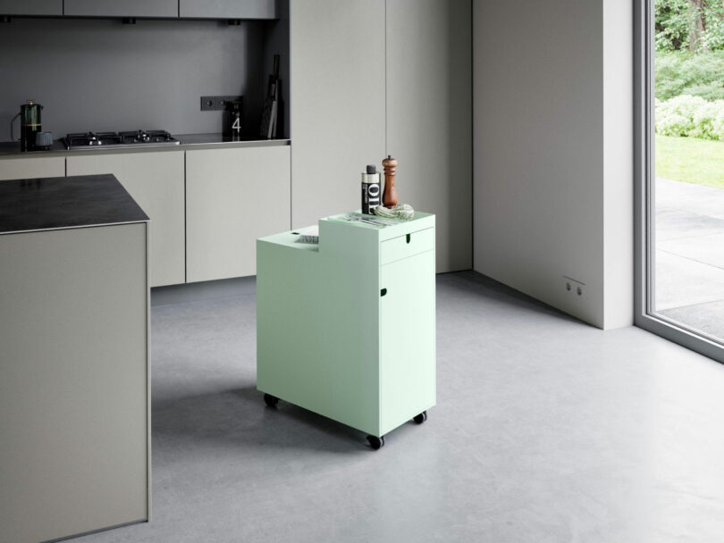 A small, mint green kitchen cart with wheels stands in a modern, minimalist kitchen near large windows. Items including pepper and salt shakers are on top.