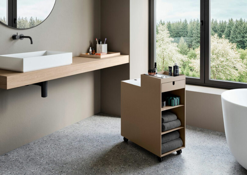 Modern bathroom with a floating wooden vanity, vessel sink, storage cart with towels and toiletries, large window, and freestanding bathtub.
