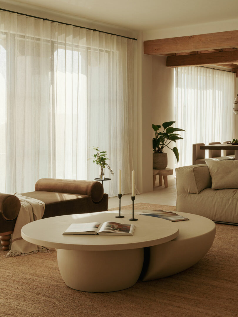Modern living room with neutral tones, which contains beige oval table, two candles, open book, pillows sitting, plants with sitting sitting, inserted curtains and clean curtains in natural light.