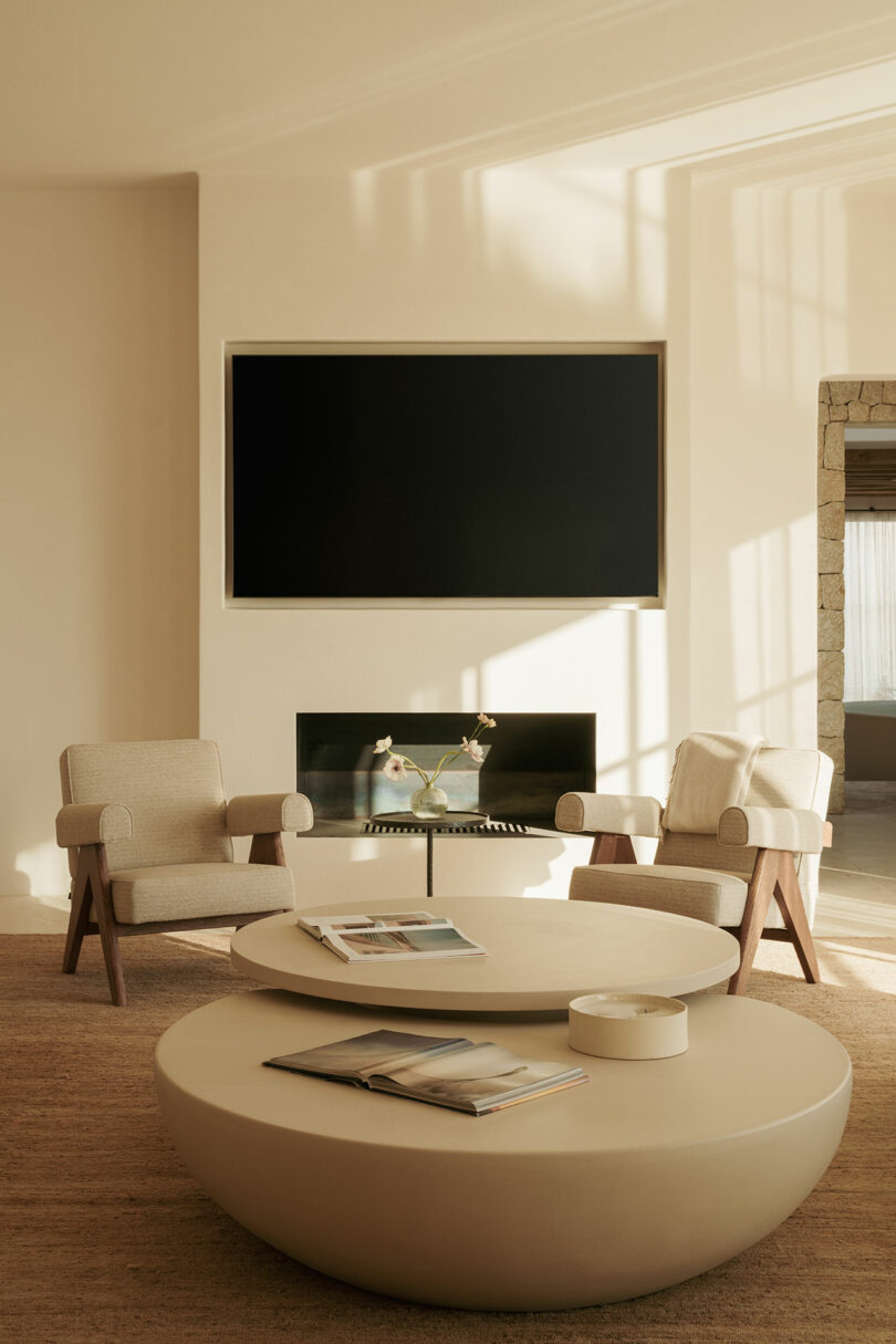 Minimalist living room with two beige armchairs, round coffee table, empty wall TV, fireplace and magazines on tables. Neutral tones and natural sunlight.