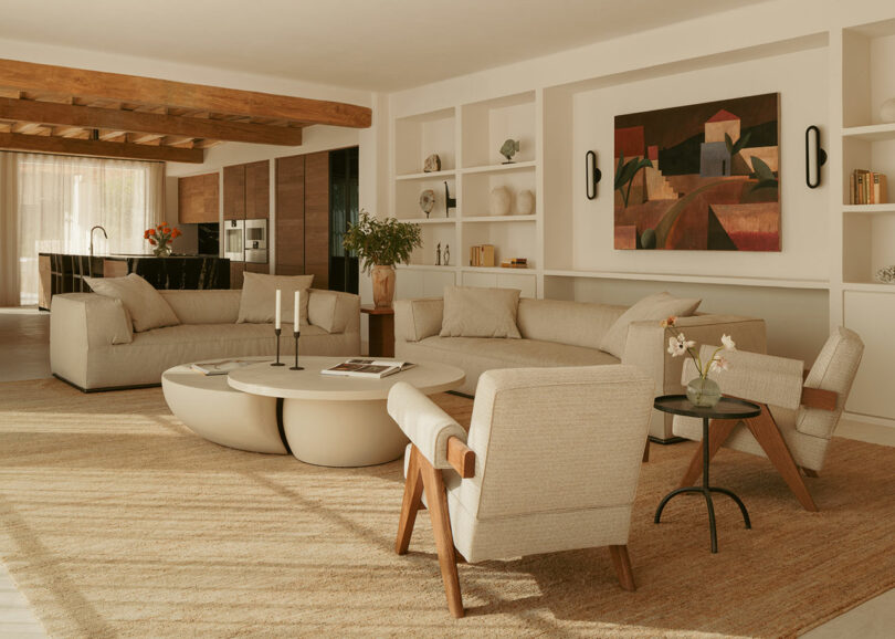 Modern living room with beige Sofi, round coffee table, wooden chairs, built-in shelves and geometric articulations on the wall, containing natural light and neutral tones.