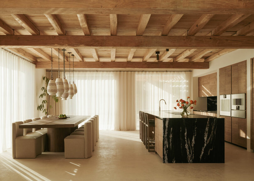 Modern kitchen and dining room with wooden ceiling, marble island, built-in devices, pendant lights and large windows with clean curtains that allow natural light.