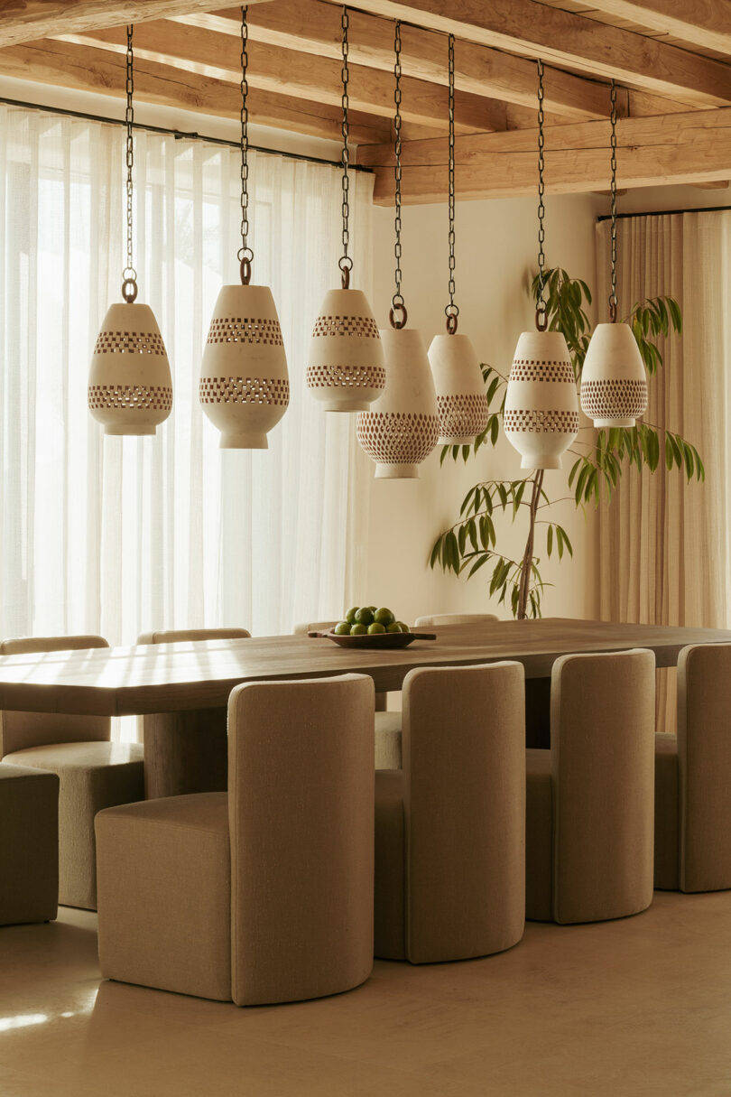 Modern dining room with a long wooden table, beige is an upholstered chair, ceramic pendant lights, clean curtains and a plant in the corner in the corner.