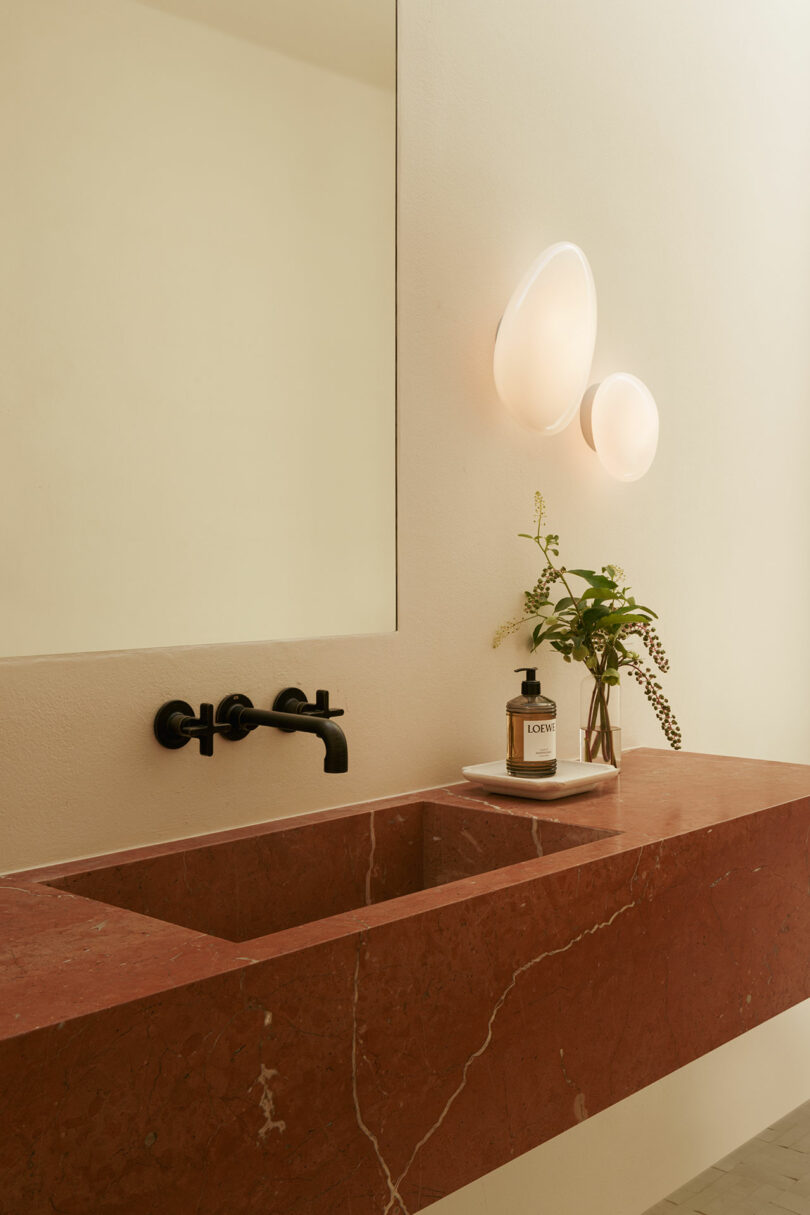 A minimalist bathroom with a red marble sink, a faucet mounted on a black wall, a large mirror, modern wall lights and small vases with greenery next to soap.
