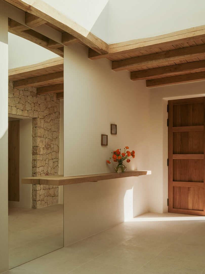 A minimalist entrance with wooden door, stone wall, wicked tiles, floating wooden shelf and glass vase of orange flowers. Two small square frames hang above the shelf.