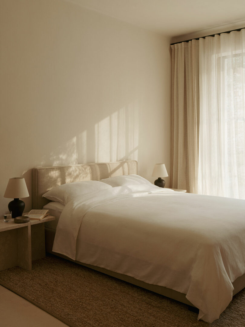 Minimalist bedroom with a neatly built bed, two nighttables with appropriate lamps, beige walls, clean curtains and soft natural light.