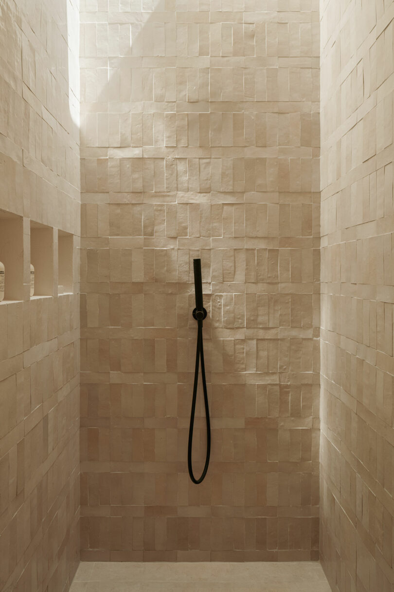 A minimalist shower with textured walls of beige tiles, black hand shower and two built-in niche shelves containing bottles.