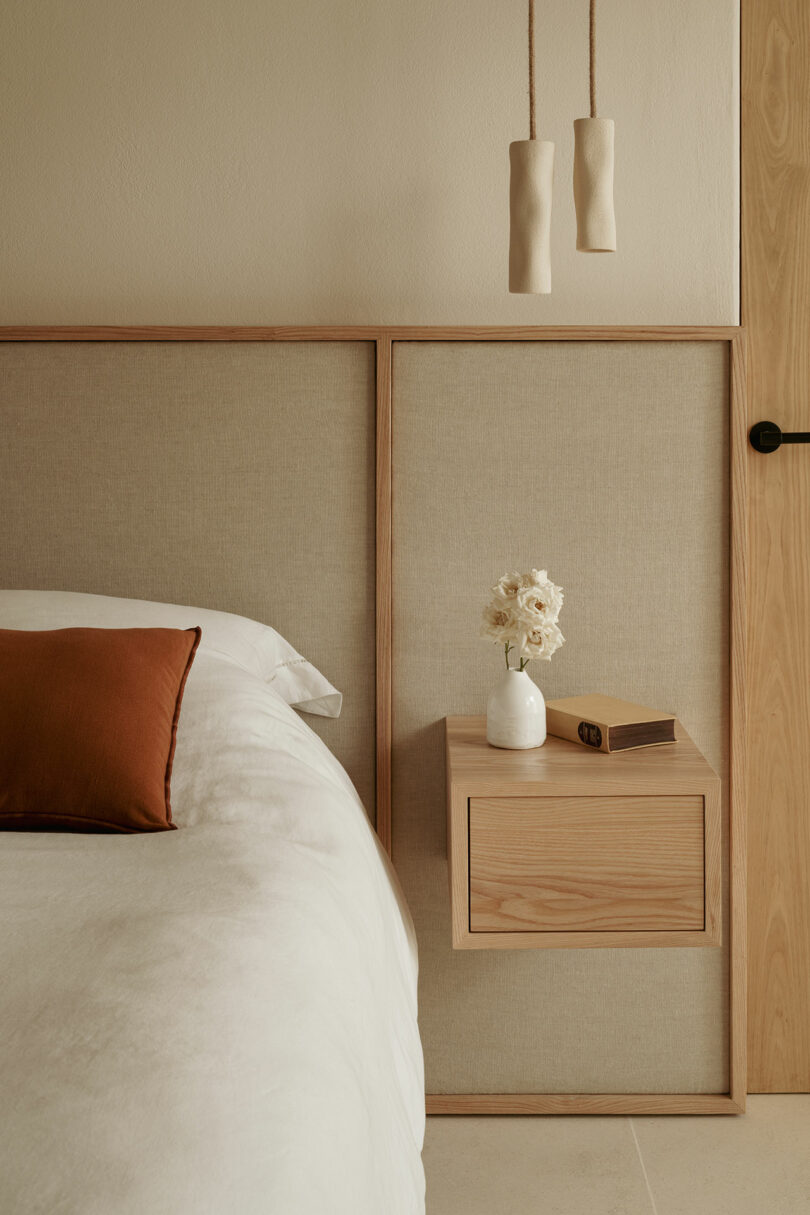 Minimalist bedroom with beige and night gender, white bedding, colored rust, small vases of white flowers, closed books and two pendants lights.