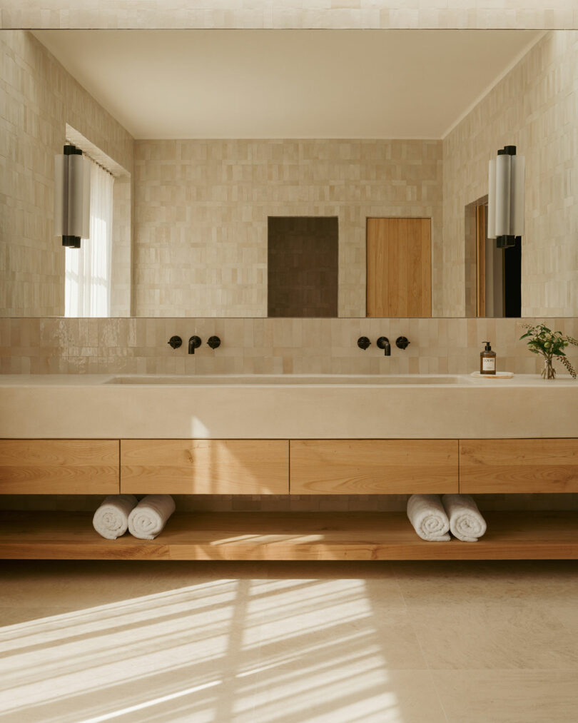 Modern bathroom with light walls tiles, large mirror, double sink with black taps, wooden vanity, rolled white towels and natural light from the window.