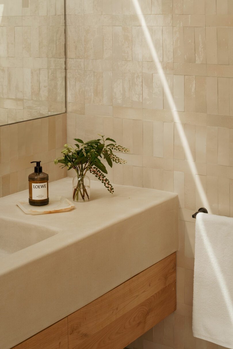 The beige paved bathroom contains stone sink wooden base, a bottle of Loeva hand soap, small vases with greenery and white towel on the rack. Sunlight Streams in.