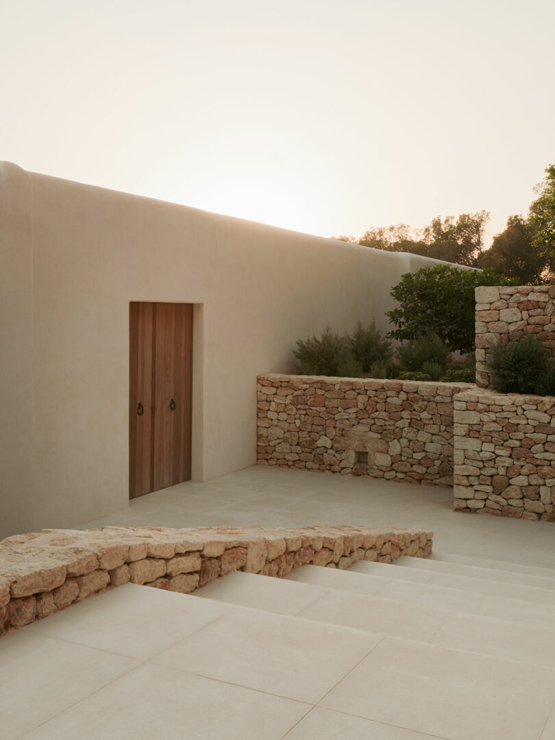 A minimalist stone staircase and walls with a wooden door, surrounded by greenery, under a warm evening sunlight.