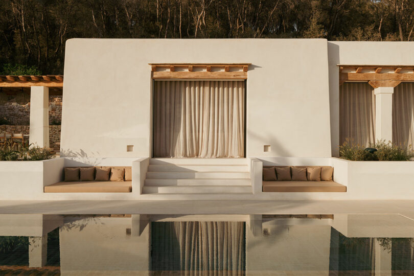 Modern white building Stucco with beige curtains, wooden accents, built-in seating and steps leading to swimming pools that reflect the structure.