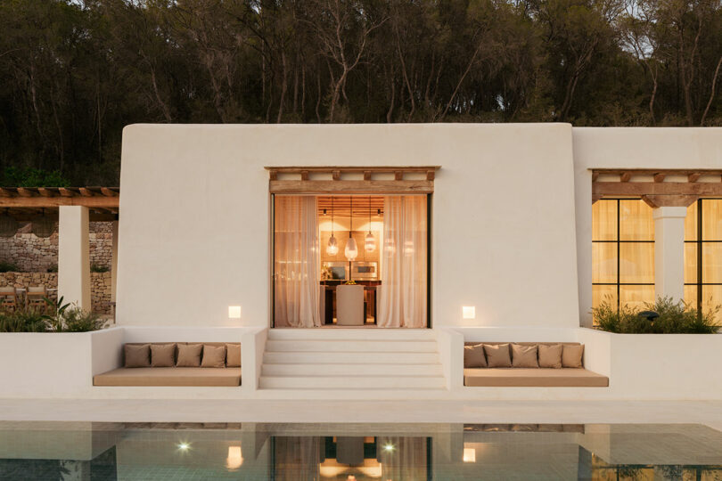Modern villa exterior with white stutter walls, symmetrical built-in sitting, large windows, central steps and reflections in the pool, placed on the background of trees.
