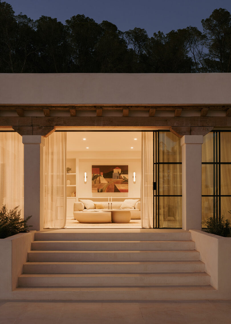 The modern, warmly lit living room is visible through a large glass door, with color light, geometric image and steps leading to the entrance to dusk at dusk.