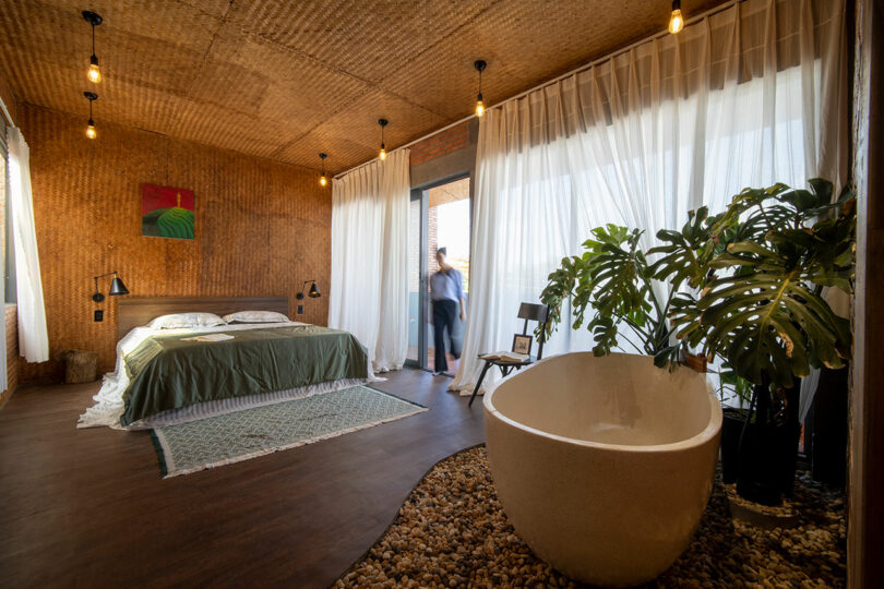 A modern bedroom with a bed, green bedding, a freestanding bathtub on pebbles, large windows with white curtains, and plants; a person stands near the balcony door.