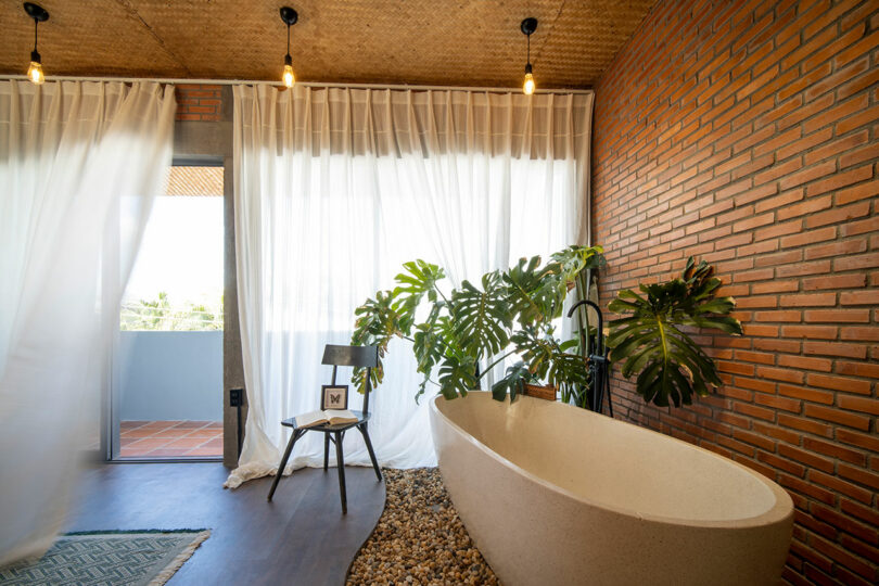 A modern bathroom with a freestanding tub, indoor plants, a wooden chair, exposed brick walls, and sheer curtains by a sliding door leading to a balcony.