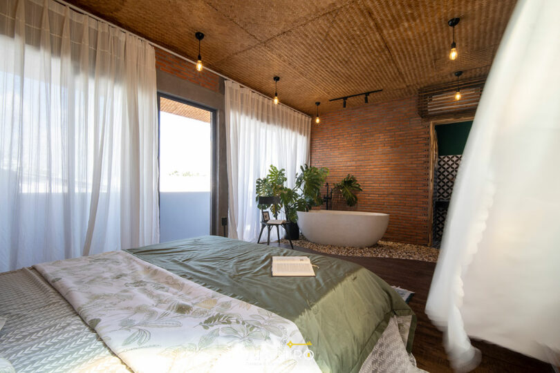 A bedroom with a large bed, open book, freestanding bathtub, potted plants, sheer curtains, and exposed brick walls under a woven ceiling with hanging light bulbs.