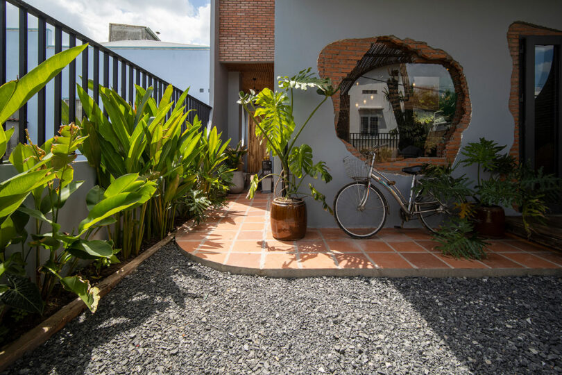 A modern patio with potted plants, a gravel walkway, and a bicycle leaning against a wall featuring a large round window with exposed brick edges.
