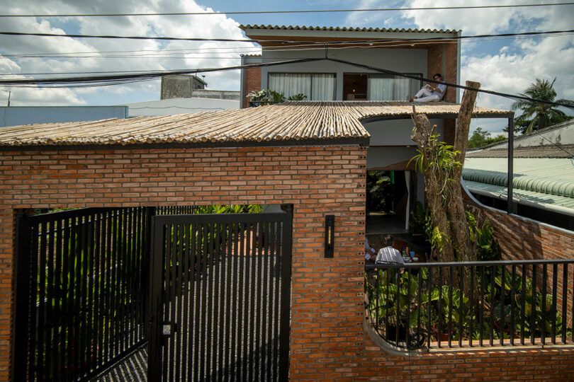 Two-story brick house with large windows and a gated entrance, featuring greenery in the front yard and two people visible, one upstairs and one downstairs.