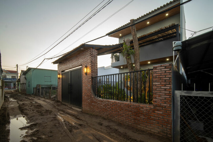 A modern house with brick and metal fencing stands beside a muddy, narrow road at dusk, with visible power lines overhead and neighboring buildings nearby.