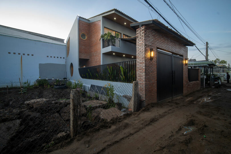 Two-story modern brick house with a black gate and balcony, exterior lights on, situated along a muddy dirt road with a fenced-off garden area.