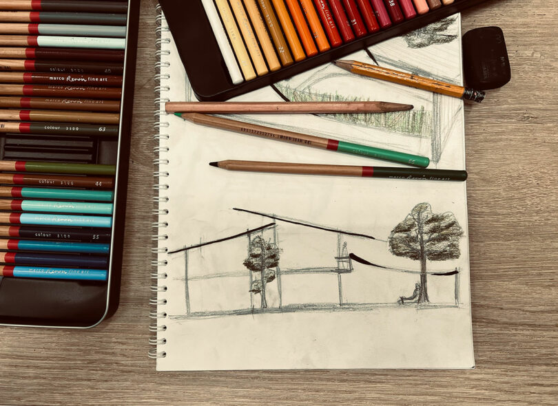 An open sketchbook shows a pencil drawing of a modern house and trees, surrounded by colored pencils and a sharpener on a wooden desk.
