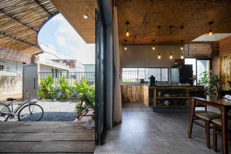 An open kitchen and dining area with wooden and bamboo decor, large windows, hanging lights, and a view of a garden with a bicycle outside.