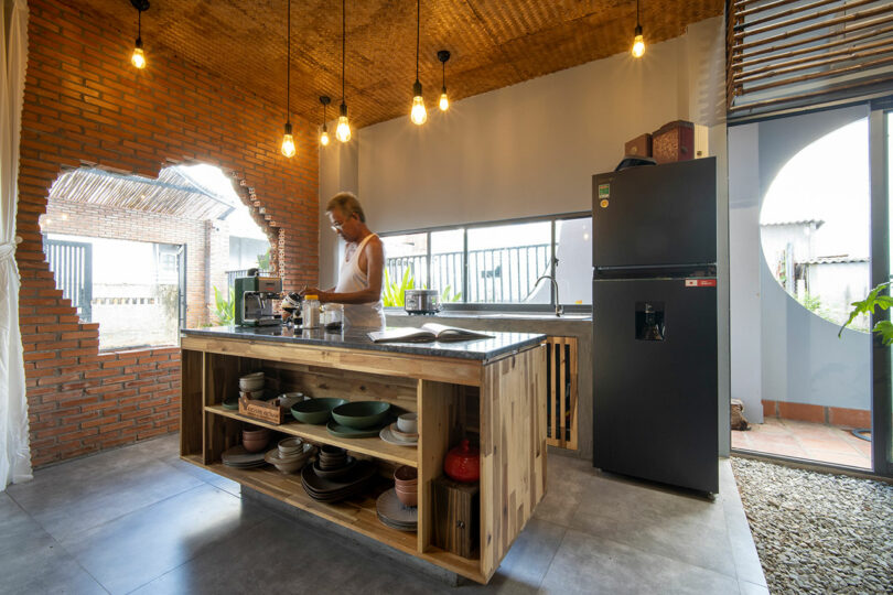 A person prepares coffee in a modern kitchen with brick walls, open shelving, pendant lights, and a large window.