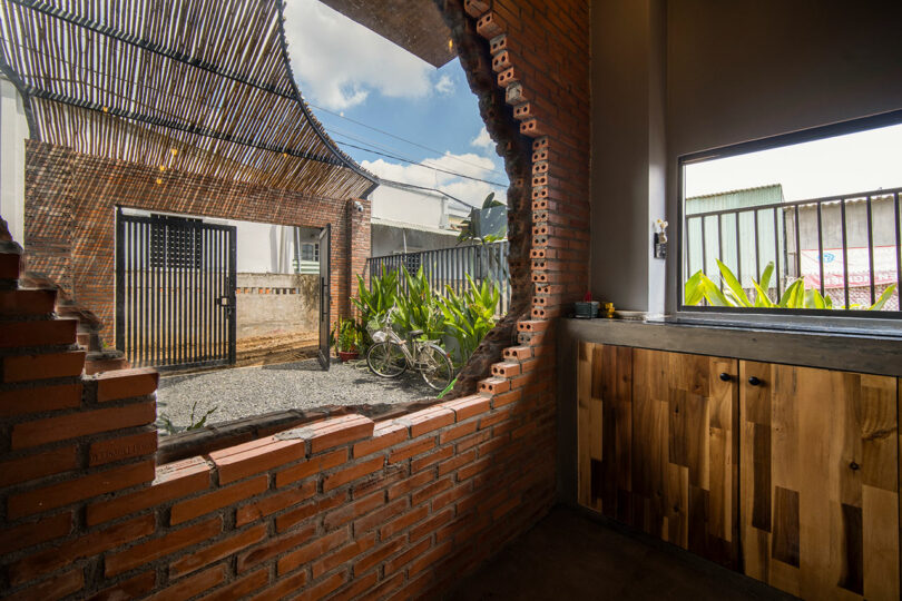 A kitchen with wooden cabinets and a stone countertop looks out through a large, irregular brick-framed window onto a courtyard with a bicycle, plants, and a striped metal gate.