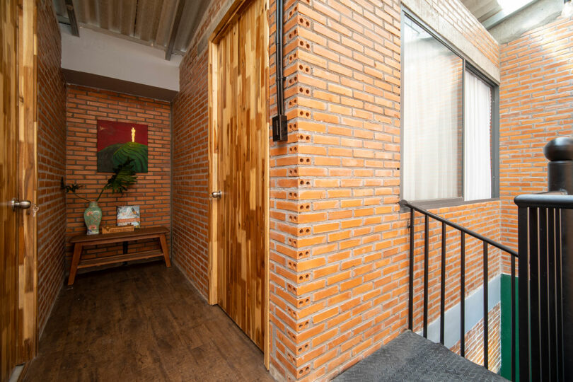 A hallway with exposed brick walls, wooden doors, a wooden bench with decorative items, and a painting, next to a black metal railing and a window.
