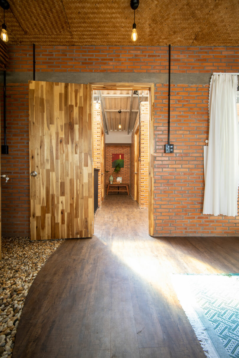 An open wooden door leads to a hallway with brick walls, a wooden floor, and a table with a potted plant at the end.