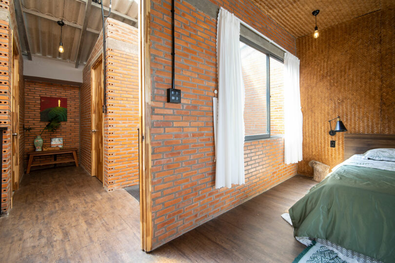 Bedroom with exposed brick walls, wooden floor, a bed with green bedding, white curtains, and an open hallway leading to another room with a plant and painting.
