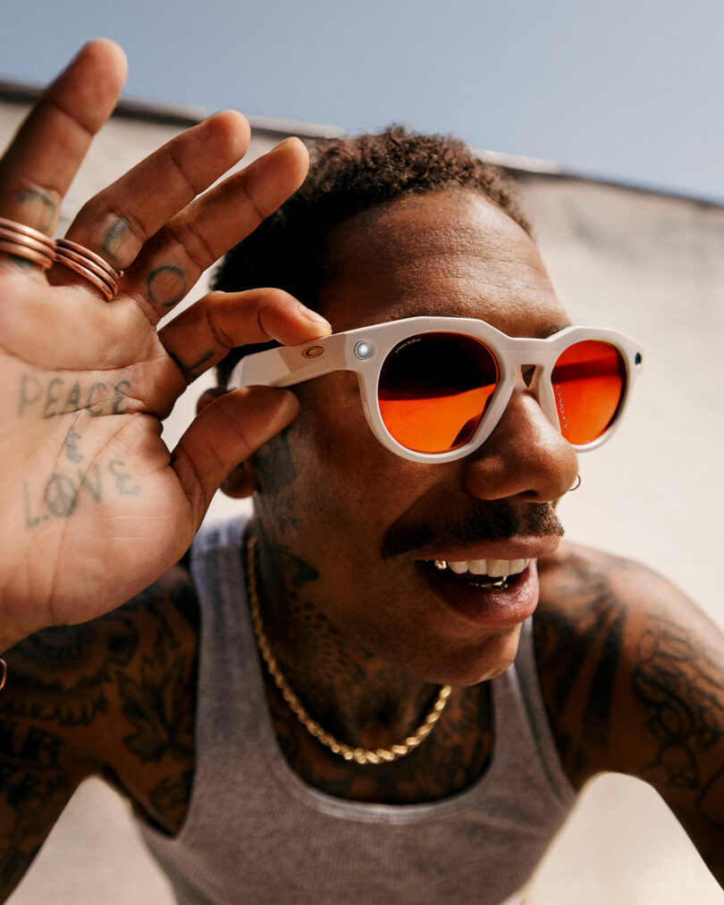 A man wearing white Meta sunglasses with orange lenses and a gray tank top smiles and holds up his hand. He has tattoos, a mustache, and wears gold chains.