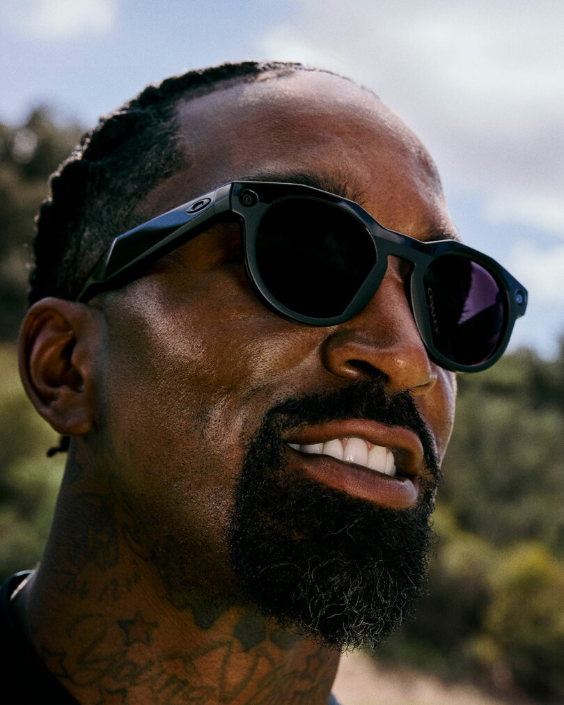 Close-up of a man with braided hair, a goatee, and neck tattoos, wearing Meta-inspired black sunglasses outdoors with a blurred background of trees and sky.